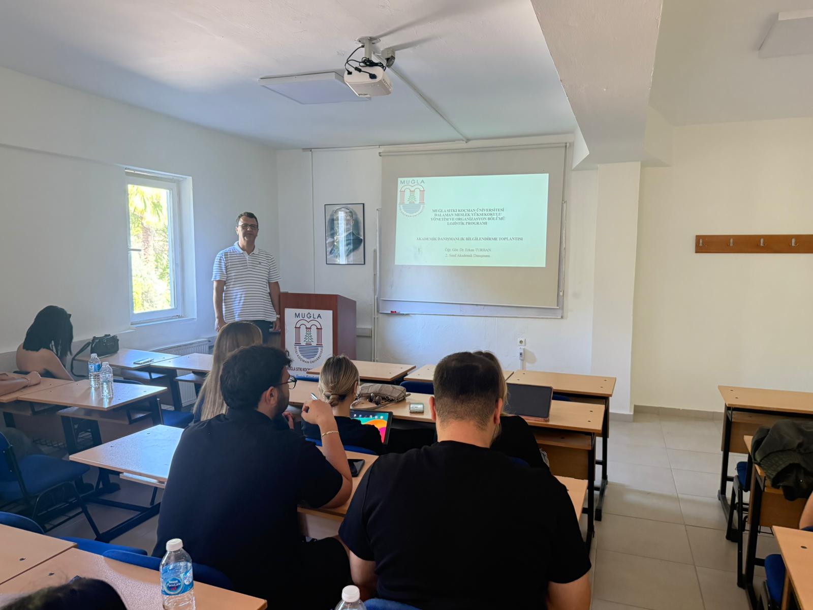 Dalaman Meslek Yüksekokulu sınıfında yapılan toplantı. Öğretim elemanı kürsüde ayakta duruyor; arkasında projeksiyon perdesine yansıtılmış sunum yer alıyor. Sınıfta sırayla oturan öğrenciler sunumu dikkatle izliyor. Pencerelerden gün ışığı içeri süzülüyor. Sunum zamanı, öğleden sonradır.